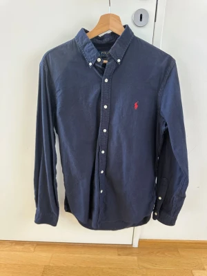 Polo Ralph Lauren skjorta  - Klassisk mörkblå skjorta från Polo Ralph Lauren med button-down krage och den ikoniska röda logotypen broderad på bröstet. Skjortan har långa ärmar, vita knappar och är tillverkad i mjuk bomull. 