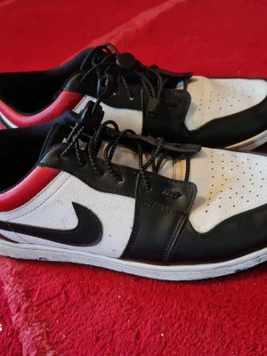 Svarta och vita Nike sneakers - Säljer ett par snygga Nike sneakers i svart, vitt och rött. Skorna har lågt skaft, klassisk swoosh-logga på sidan och ovandel i läder med perforerade detaljer på tån. Utrustade med svarta snören och bekväm sula. Perfekta för dig som gillar streetwear.