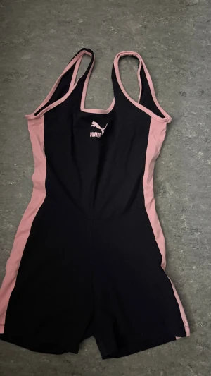 Svart och rosa träningsdräkt Puma - Säljer en svart och rosa träningsdräkt från Puma med smala axelband och logga på bröstet. Dräkten har en sportig design med rosa paneler längs sidorna och runt kanterna. Perfekt för gym, dans eller annan träning.