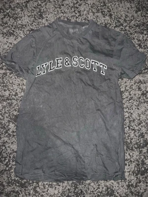 Grå Lyle & Scott t-shirt - Snygg grå t-shirt från Lyle & Scott med vit logga tryckt över bröstet. Klassisk passform och rund halsringning. Perfekt till jeans eller shorts för en enkel och stilren look.