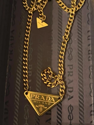 Statement-halsband, made in Italy, ikonisk logotyp  - Mycket snyggt och coolt halsband i guldfärgad metall med en ikonisk triangelformad logotyp: ”PRADA MILANO”. Kedjan är grov och har även en liten triangelberlock med logotyp. Perfekt för dig som vill ha en unik statement-accessoar. Ej verifierad Prada, därav det låga priset. Passa på!