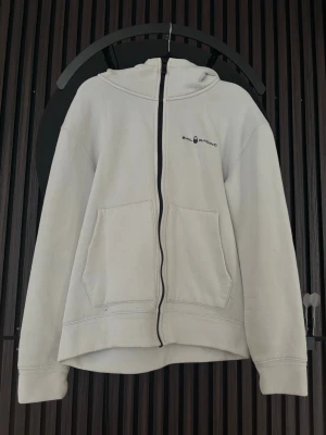 Vit hoodie från Sail Racing - Vit hoodie från Sail Racing med dragkedja framtill och två stora fickor. Lite nopprig samt har en fläck på luvan.