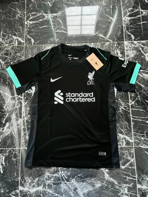 Svart Liverpool Nike matchtröja - Snygg svart Liverpool FC matchtröja från Nike med vita loggor och detaljer i turkos på ärmarna. Tröjan har rund hals, korta ärmar och är tillverkad i ett lätt, ventilerande funktionsmaterial. Perfekt för fotboll eller träning.