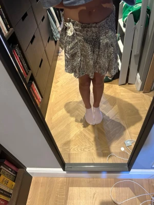 Kort omlottkjol med volang och knyt - Säljer en kort omlottkjol med volangkant och knyt i midjan. Kjolen har ett småmönstrat tryck i beige, brunt och grönt och är gjord i ett lätt och luftigt material. Perfekt för dig som gillar en avslappnad men trendig stil.