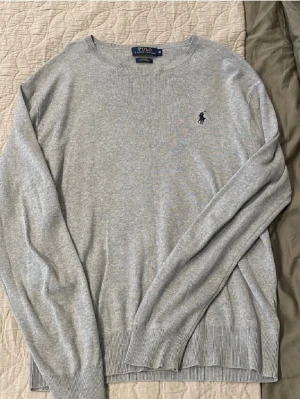 Ralph lauren stickat - Snygg ljusgrå stickad tröja från Polo Ralph Lauren med klassisk rund halsringning och mörkblå broderad logga på bröstet. Tröjan har ribbade muddar vid ärmslut och nederkant. Perfekt till lager på lager och enkel att matcha.
