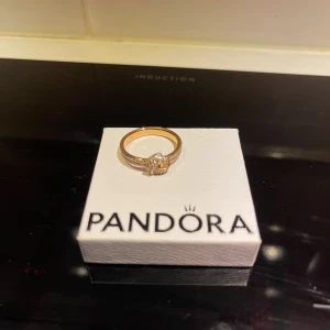 Ring i roséguld med stenar från Pandora - Snygg roséuldfärgad ring från Pandora med glittriga stenar och en elegant knutdesign framtill. Storlek 60 (60 mm).