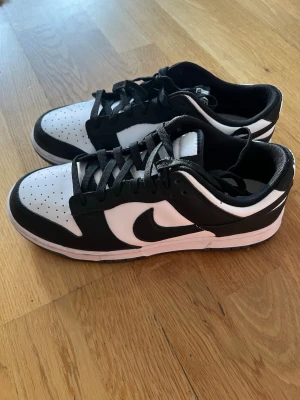 Nike Dunk Low svart/vit sneakers - Säljer ett par Nike Dunk Low sneakers i svart och vitt. Skorna har klassisk låg siluett, svart swoosh och vita partier med perforerad tåbox. Tillverkade i läder med svart snörning och vit sula. Perfekta för dig som gillar stilrena och tidlösa sneakers.