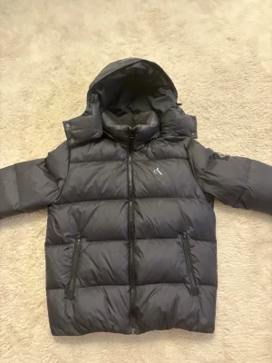 Svart pufferjacka från Calvin Klein - Snygg svart pufferjacka från Calvin Klein Jeans med huva och dragkedja framtill. Jackan har två framfickor med dragkedja och en tydlig CK-logga på ärmen. Perfekt för kalla vinterdagar och riktigt trendig look. 