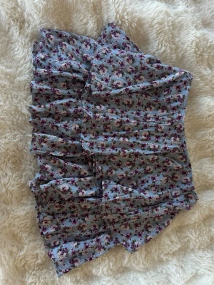 Blommig volangkjol från Zara - Supersöt kort kjol från Zara i ljusblått med blommigt mönster i lila och beige toner. Kjolen har volangdetaljer och omlottkänsla som ger en flowy och feminin look. Perfekt för dig som gillar romantisk stil!