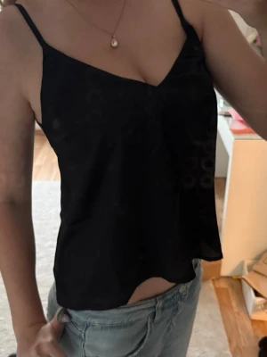 Svart linnetopp med smala axelband - Säljer en svart linnetopp med smala axelband och v-ringad hals. Toppen har en slät, glansig yta som ger en stilren look och är perfekt till jeans eller kjol. 