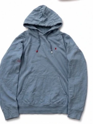 Blå hoodie med broderad detalj - Säljer en ljusblå hoodie med huva och dragsnören med röda detaljer. Tröjan har en känguruficka framtill och en liten röd broderad detalj på bröstet. Muddar vid ärmslut och nederkant. Perfekt för en chill och avslappnad stil.