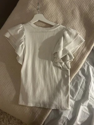 Vit ribbad topp med volangärmar - Supersnygg vit topp i ribbat material med breda volangärmar som ger en cool och trendig look. Rak passform och rund halsringning.