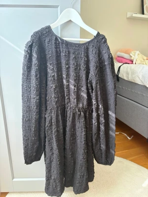 Svart strukturerad långärmad klänning - Säljer en svart kortklänning med lång ärm och coolt strukturerat tyg. Klänningen har rund halsringning och ett lätt puffigt fall. Perfekt för dig som gillar unika detaljer och en loose fit.