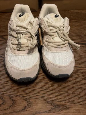 Beige Acics gel - Snygga sneakers från Asics i beige med detaljer i mesh och mocka. Skorna har en chunky grå sula och klassisk snörning. Perfekta för en sportig och trendig look.
