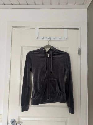 Mjuk grå velourhoodie med dragkedja - Säljer en grå hoodie i mjuk velour med dragkedja framtill och snörning i huvan. Hoodien har långa ärmar, kängurufickor och ribbade muddar. Perfekt för en chill och avslappnad stil. Dock har den ena metallbiten från snöret tappats