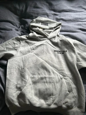 Grå Essentials Fear of God hoodie - Snygg ljusgrå hoodie från Essentials Fear of God med svart logga på bröstet och patch på ärmen. Klassisk känguruficka framtill och stor huva. Tillverkad i mjukt bomullsmaterial som är perfekt för chill dagar. Enkel och clean design som passar till allt.