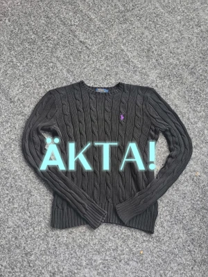 Buda på en äkta Svart kabelstickad tröja Polo Ralph Lauren - Kabelstickad svart tröja från Polo Ralph Lauren med lila broderad logga på bröstet. Klassisk rund halsringning och ribbade muddar vid ärmslut och nederkant. Perfekt för dig som gillar stilrena och tidlösa plagg.