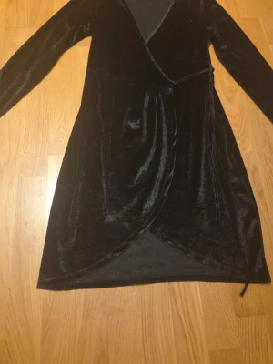 Svart omlottklänning i sammet - Snygg svart omlottklänning i mjuk sammet med lång ärm. Klänningen har en omlottdesign framtill och är knälång, vilket ger en elegant och trendig look. Perfekt för dig som vill ha något stilrent och bekvämt.