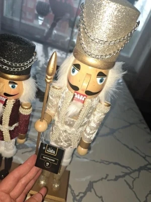 Två dekorativa nötknäppare - Säljer två klassiska nötknäppare i trä, en i vinrött och guld med svart hatt och en i vitt och guld med glittrig hatt. Båda har detaljer som kedjor, mustasch och håller varsin stav. Perfekta som juldekoration eller samlarpryl.