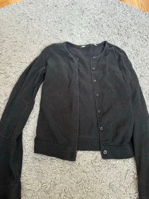 Svart basic kofta med rund hals från H&M - En enkel svart kofta med rund halsringning och knäppning framtill från H&M. Långa ärmar och ribbade muddar vid ärmslut och nederkant. Perfekt att slänga över en topp eller linne för en clean look.