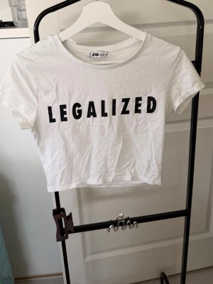 Vit croppad t-shirt med tryck - Säljer en vit croppad t-shirt med svart text 'LEGALIZED' på bröstet. T-shirten har korta ärmar och rund halsringning. Perfekt för dig som gillar statement-plagg och enkel stil.