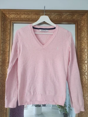 Ljusrosa stickad tröja Polo Club - Snygg ljusrosa stickad tröja från Polo Club med V-ringning och diskret broderad logga på bröstet. Tröjan har långa ärmar och ribbade muddar vid ärmslut och nederkant. Perfekt att matcha med jeans eller kjol för en clean look.