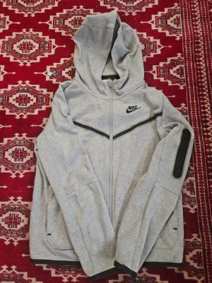 Grå hoodie från Nike med dragkedja - Snygg grå hoodie från Nike med hel dragkedja och svart Nike-logga på bröstet. Hoodien har svarta detaljer, fickor på sidorna och en extra ficka med dragkedja på ärmen. Tillverkad i mjukt material och har en klassisk huva. Bara att fråga om bilder. Pris kan så klart diskuteras😉