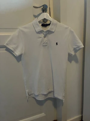 Vit pikétröja från Polo Ralph Lauren - Klassisk vit pikétröja från Polo Ralph Lauren med korta ärmar och krage. Tröjan har den ikoniska broderade loggan på bröstet och knäppning vid halsen. Perfekt till jeans eller shorts för en clean och stilren look. Storleken är junior L (14-16) och slimfit. Inköpt på Kidsbrandstore. 
