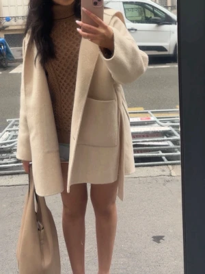 Beige kappa i ull - Stilren beige kappa med huva och knytskärp i midjan. Kappan har raka ärmar, stora utanpåfickor och en avslappnad passform. Perfekt för lager på lager och enkel att matcha till olika outfits. Materialet ser mjukt och lite ulligt ut. (Ny pris över 3 tusen)