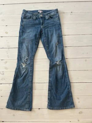 Blå bootcut jeans med slitningar - Säljer ett par blå bootcut jeans med slitningar på båda benen och snygg broderad ficka bak med silverdetaljer. Jeansen har klassisk femficksmodell och låg midja. Perfekta för dig som gillar en avslappnad men trendig stil.