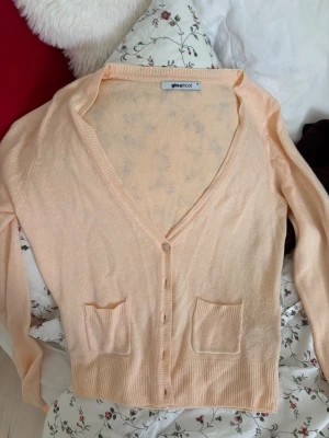 Persikofärgad kofta från Gina Tricot - Söt persikofärgad kofta från Gina Tricot med v-ringning, knäppning framtill och två små fickor. Koftan har långa ärmar och ribbade muddar. Broderad detalj på bröstet. Perfekt för lager på lager och en mjuk känsla.