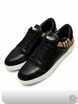 Svarta sneakers från burburry - Snygga svarta sneakers med klassiskt rutigt mönster på hälen och vita sulor. Skorna har svarta snören och detaljer i mocka och läder. Perfekta för dig som gillar en stilren men ändå trendig look. Köpta från burberry äkthets bevis kommer med.