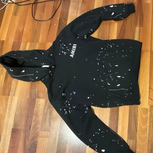 Svart hoodie från Amiri med färgstänk - Säljer en svart hoodie från Amiri med vita och grå färgstänk över hela plagget. Hoodien har huva, magficka och ribbade muddar vid ärmslut och nederkant. Amiri-loggan syns på bröstet. Perfekt för dig som gillar streetwear och unik design.