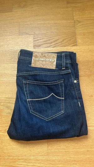 Mörkblå jeans från Jacob Cohen - Snygga mörkblå jeans från Jacob Cohen med kontrastsömmar och klassisk femficksmodell. Jeansen har en patch i mocka baktill och detaljerade knappar framtill. Tillverkade i mjukt denimtyg med exklusiv känsla och diskret broderad logga på fickan. Jeansen är i modell Bard och använda en handfull gånger, super fint skick!
