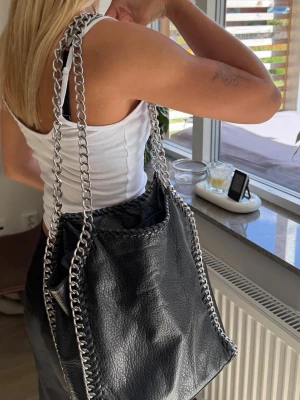 Svart axelväska med kedjedetaljer - Snygg svart axelväska i fejkläder med grova silverfärgade kedjor som axelband och längs kanterna. Väskan har en mjuk, slouchy form och en modern, trendig look som passar perfekt till streetstyle och casual outfits.