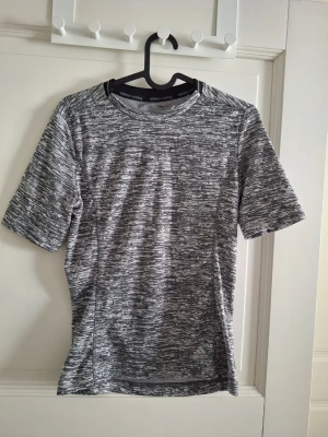 Gråmelerad Adidas träningströja - Gråmelerad träningströja från Adidas med korta ärmar och rund hals. Tillverkad i ett lätt funktionsmaterial som andas. Diskret Adidas-logga framtill och tre klassiska ränder på ryggen för en sportig look.