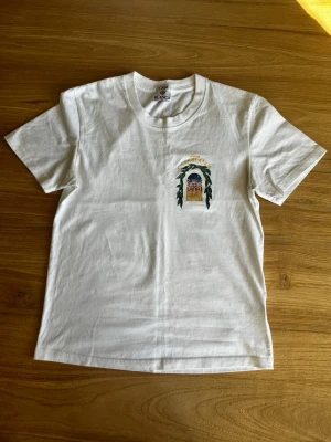 Vit t-shirt från Casablanca - Vit t-shirt från Casablanca med tryck på bröstet som föreställer en byggnad och texten 'Avenida Casablanca'. T-shirten har rund hals och korta ärmar. Tillverkad i mjuk bomull och har en clean, stilren look med en liten färgklick från trycket.