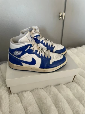 Air Jordan 1 Mid blå/vit - Orginal priset är ungefär 1000 upp till 2400. Säljer ett par Air Jordan 1 Mid sneakers i blått och vitt läder. Klassisk siluett med blå swoosh, vita snören och Air Jordan-logga på sidan. Skorna har perforerad tåbox och vadderad krage för extra komfort. Perfekta för dig som gillar retrostil. 