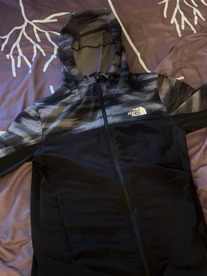 Svart vindjacka från The North Face - Snygg svart vindjacka från The North Face med grått och vitt mönster upptill och på huvan. Jackan har hel dragkedja framtill, huva och logga på bröstet. Perfekt för blåsiga dagar och enkel att matcha med olika outfits.