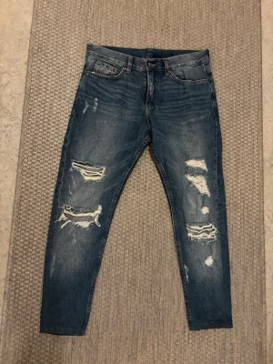 Blå slitna slim jeans från H&M Divided - Säljer ett par blå slim fit jeans från H&M Divided med snygga slitningar och hål på båda benen. Jeansen har klassisk femficksmodell och är tillverkade i bomull med lite stretch för skön passform. Perfekta för en trendig och avslappnad look.