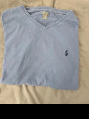 Blå v-ringad t-shirt från Polo Ralph Lauren - Säljer en stilren blå t-shirt från Polo Ralph Lauren med v-ringning och den klassiska broderade loggan på bröstet. T-shirten är i mjuk bomull och har en normal passform. Perfekt till jeans eller shorts för en clean look.