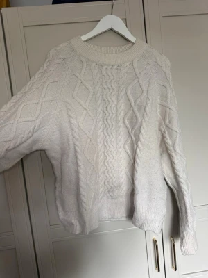 Vit kabelstickad tröja - Kabelstickad tröja i mjuk vit ton med klassiskt flätmönster framtill och på ärmarna. Tröjan har rund halsringning, ribbade muddar och avslut. Perfekt för lager på lager och riktigt cozy vibes. Kan finnas skavanker eller liknande 
