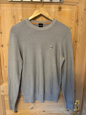 Ljusgrå ulltröja från HUGO BOSS - Snygg ljusgrå tröja från BOSS med rund halsringning och diskret logga på bröstet. Tröjan har långa ärmar och ribbade muddar vid ärmslut och nederkant. Perfekt till jeans eller chinos för en clean look. 100% ull                                                     Storlek M men passar mer som en S.