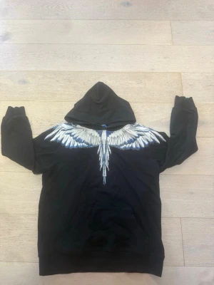 Svart hoodie med vita vingar - Säljer en svart hoodie med stor huva och ett fett vingmotiv i vitt och blått över axlarna, både fram och bak. Hoodien har långa ärmar och ribbade muddar. Perfekt för dig som vill sticka ut med en unik design.