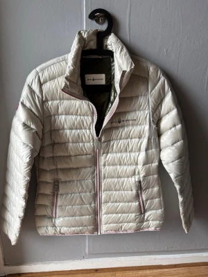 Ljusbeige dunjacka från Sail Racing - Snygg ljusbeige dunjacka från Sail Racing med quiltad design och två dragkedjeförsedda fickor framtill. Jackan har hög krage, diskret logga på bröstet och kontrastfärgade detaljer vid dragkedjorna. Perfekt för kalla vinterdagar.