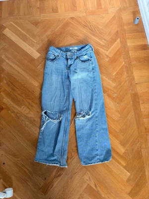 Ljusblå slitna baggy jeans från Levi  - Säljer ett par ljusblå jeans med raka ben och stora slitningar vid knäna. Jeansen har fransiga benslut och klassisk femficksmodell. Materialet är denim i bomull och passformen är avslappnad och trendig. Den är lite söndriga på botten.