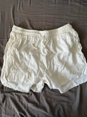 Vita linneshorts med snörning - Säljer ett par vita shorts i linne med elastisk midja och snörning framtill. Shortsen har avslappnad passform och sidofickor, perfekta för varma dagar. Luftiga och sköna att bära till sommaren.