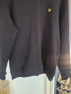 Svart sweatshirt från Lyle & Scott - Klassisk svart sweatshirt från Lyle & Scott med gul broderad logga på bröstet. Tillverkad i ekologisk bomull och har ribbade muddar vid ärmslut och nederkant. Enkel och stilren design som passar till det mesta.
