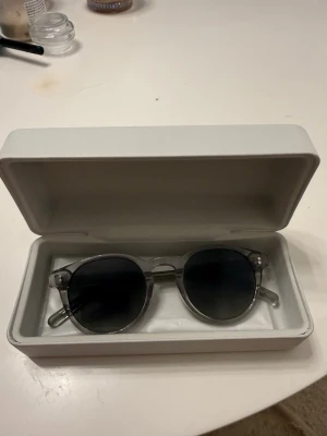 CHIMI 03 Gray - Säljer ett par stilrena solglasögon med rund form och transparent båge. Glasen är mörka och ger en cool kontrast till den klara ramen. Perfekta för dig som vill ha en modern och trendig look i sommar. Kommer med ett vitt hårt fodral.