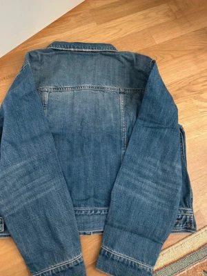Klassisk blå jeansjacka från H&M - Snygg blå jeansjacka från H&M med raka ärmar, silverfärgade knappar och två bröstfickor med lock. Jackan har en klassisk krage och är gjord i slitstarkt denimtyg. Perfekt för dig som gillar en tidlös och avslappnad stil.